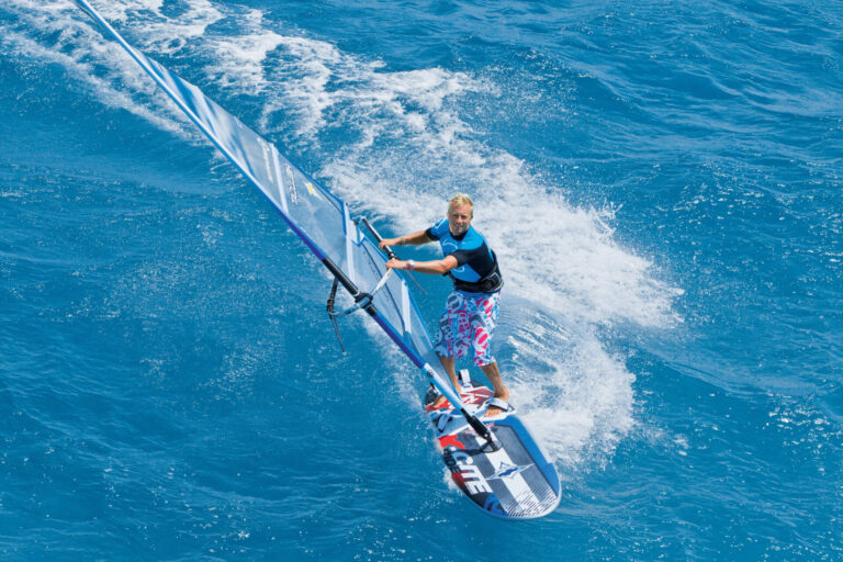 En person, der windsurfer på klart blåt vand, balancerer på et bræt, mens han holder fast i et sejl. Personen er iført en sort og blå våddragt og farverige shorts, hvilket skaber en livlig scene på baggrund af det pulserende hav.