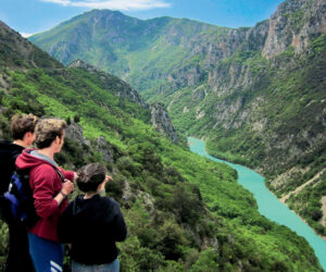 Tre personer står på en klippe med udsigt over en grøn flod, der snor sig gennem en frodig dal, omgivet af bjerge under en delvist overskyet himmel.