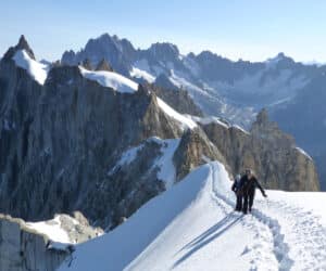 To klatrere, der går langs en snedækket bjergryg med tårnhøje klippetoppe i baggrunden. Himlen er klar og blå og fremhæver det dramatiske og barske landskab.
