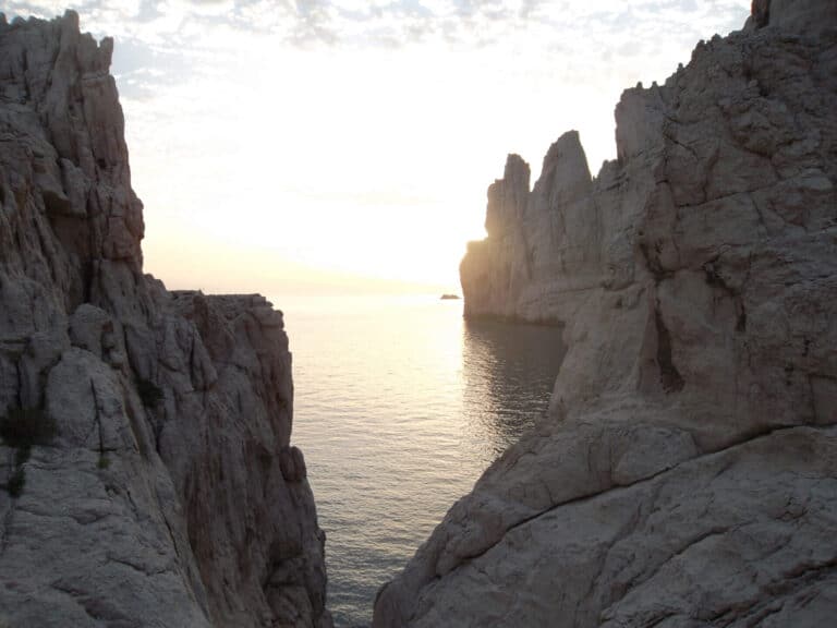 Solnedgangsudsigt over et roligt hav indrammet af forrevne kalkstensklipper, der skaber en smal passage. Himlen har en blød gradient af farver, og klippeformationerne tilføjer dramatisk kontrast til det fredfyldte vand.
