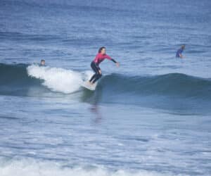 En surfer i en pink top rider på en lille bølge på et surfbræt med to andre surfere i baggrunden på et roligt hav.