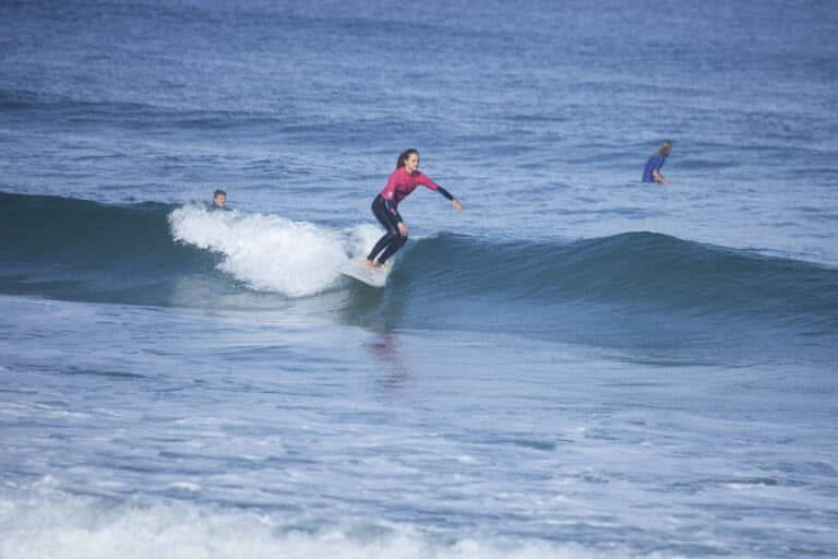 En surfer i en pink top rider på en lille bølge på et surfbræt med to andre surfere i baggrunden på et roligt hav.