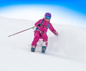 En skiløber i lyserødt gear og en lilla hjelm kører ned ad en sneklædt skråning og skaber et spor af pudder i luften. Den klare blå himmel danner baggrunden og fremhæver hastigheden og spændingen ved nedstigningen.