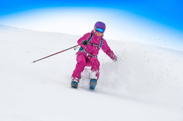 En skiløber i lyserødt gear og en lilla hjelm kører ned ad en sneklædt skråning og skaber et spor af pudder i luften. Den klare blå himmel danner baggrunden og fremhæver hastigheden og spændingen ved nedstigningen.