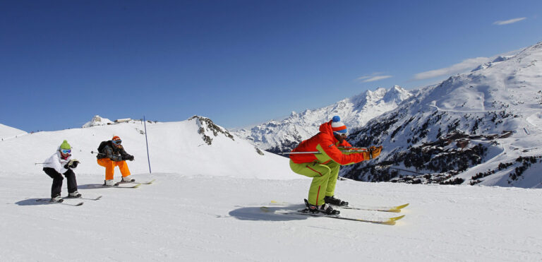Tre personer på ski ned ad bakke på en solrig vinterdag. Skiløberne er iført farverige outfits: orange, grøn og hvid, med sneklædte bjerge i baggrunden under en klar blå himmel.