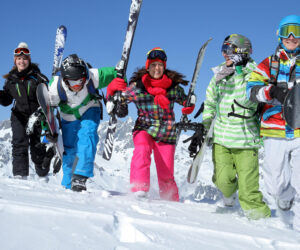 En gruppe på fem personer, klædt i farverigt skiudstyr, går gennem sneen med ski og snowboards. De smiler og ser ud til at nyde en solskinsdag i et sneklædt bjerglandskab.