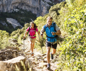 To mennesker, der vandrer energisk på en stenet sti i et bjergrigt landskab. Begge bærer vandreudstyr, herunder rygsække og bruger vandrestave. Baggrunden har grønt løv og et stort klippefyldt bjerg under en klar himmel.