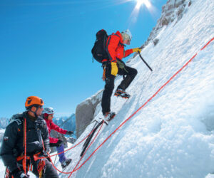 Tre klatrere, udstyret med hjelme og klatreudstyr, bestiger en stejl, sneklædt bjergside under en klar blå himmel. De er forbundet med reb, hvor en klatrer leder og andre følger tæt. Solen skinner klart ovenover.