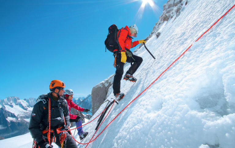 Tre klatrere, udstyret med hjelme og klatreudstyr, bestiger en stejl, sneklædt bjergside under en klar blå himmel. De er forbundet med reb, hvor en klatrer leder og andre følger tæt. Solen skinner klart ovenover.