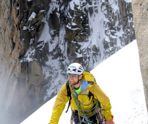 En klatrer i gul jakke og hjelm bestiger en sneklædt bjergskråning ved hjælp af en isøkse som støtte. Klippeflader dækket af sne er synlige i baggrunden. Klatreren har rygsæk og klatreudstyr på.
