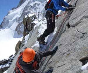 Tre klatrere bestiger en stejl, sneklædt klippeflade. Den ledende klatrer er iført blåt og bruger reb til støtte, mens en anden klatrer med orange hjelm følger efter. En tredje klatrer observerer bagfra. En snedækket bjergtop er synlig i baggrunden.