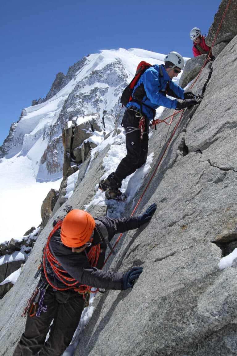 Tre klatrere bestiger en stejl, sneklædt klippeflade. Den ledende klatrer er iført blåt og bruger reb til støtte, mens en anden klatrer med orange hjelm følger efter. En tredje klatrer observerer bagfra. En snedækket bjergtop er synlig i baggrunden.