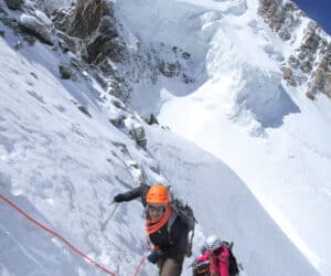 To klatrere bestiger en stejl, sneklædt bjergskråning. Den førende klatrer i en orange hjelm griber et reb, tæt fulgt af en anden i pink gear. Skarvede tinder og en klar blå himmel indrammer scenen.