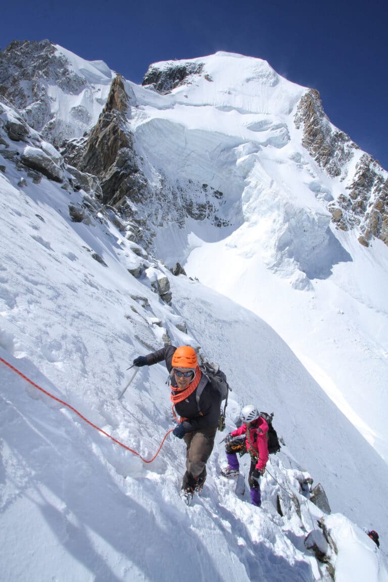 To klatrere bestiger en stejl, sneklædt bjergskråning. Den førende klatrer i en orange hjelm griber et reb, tæt fulgt af en anden i pink gear. Skarvede tinder og en klar blå himmel indrammer scenen.