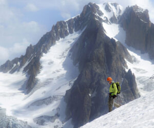 To klatrere i lyse jakker krydser en sneklædt bjergryg, trukket sammen for sikkerheds skyld. Majestætiske, takkede tinder rejser sig i baggrunden mod en delvist overskyet himmel.