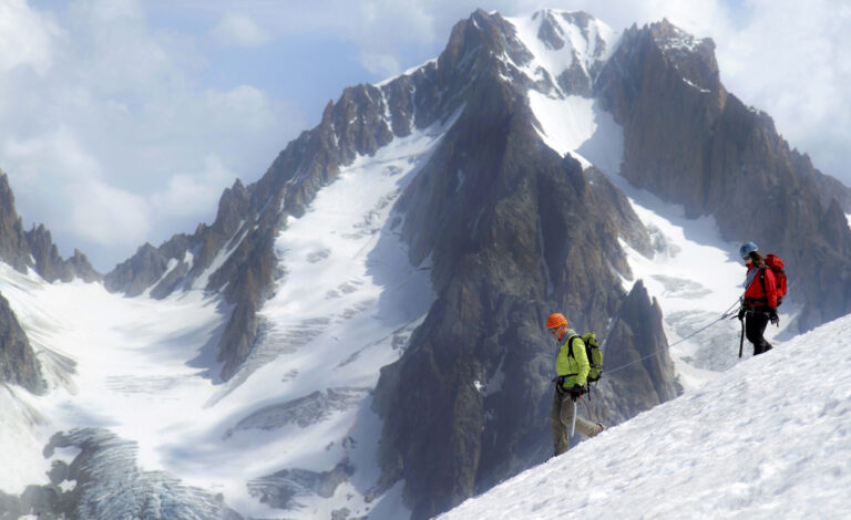 To klatrere i lyse jakker krydser en sneklædt bjergryg, trukket sammen for sikkerheds skyld. Majestætiske, takkede tinder rejser sig i baggrunden mod en delvist overskyet himmel.