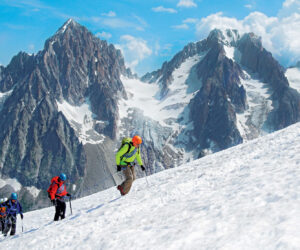 En gruppe klatrere bestiger en sneklædt bjergskråning under en klar blå himmel. Skarvede, snedækkede tinder rager i baggrunden og viser et dramatisk alpelandskab.