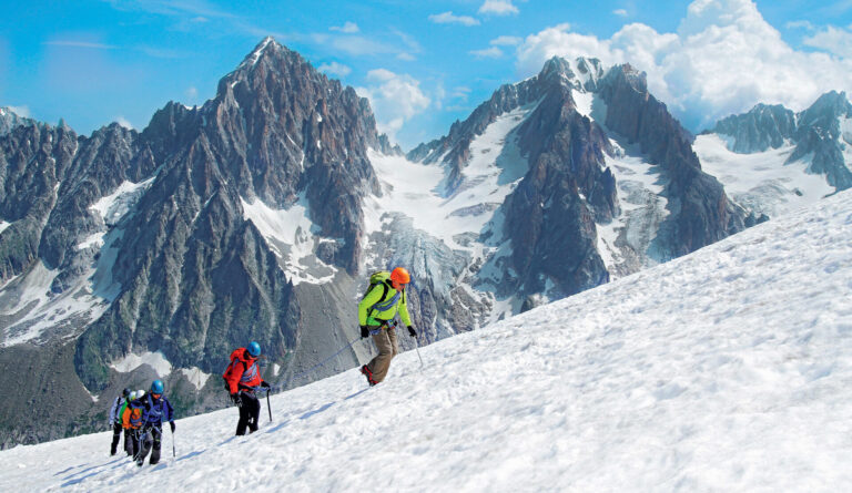 En gruppe klatrere bestiger en sneklædt bjergskråning under en klar blå himmel. Skarvede, snedækkede tinder rager i baggrunden og viser et dramatisk alpelandskab.