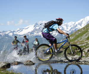 En gruppe mountainbikere kører gennem en lavvandet å på en stenet sti. Den førende cyklist skaber et plask, mens andre følger tæt. Sneklædte bjerge, der minder om et skiferieeventyr, og en klar blå himmel danner en fantastisk kulisse.