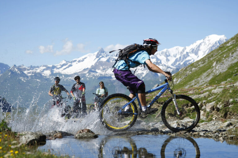 En gruppe mountainbikere kører gennem en lavvandet å på en stenet sti. Den førende cyklist skaber et plask, mens andre følger tæt. Sneklædte bjerge, der minder om et skiferieeventyr, og en klar blå himmel danner en fantastisk kulisse.
