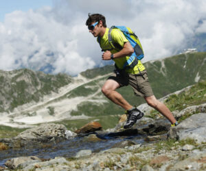 En mand i gul skjorte og kakishorts med blå rygsæk løber på stenet terræn i bjergene. Han har solbriller på og springer over et lille vandløb med en overskyet himmel og tinder i baggrunden.