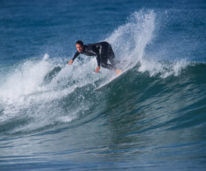 En surfer i en sort våddragt rider dygtigt på en bølge og skærer sig gennem vandet med en dynamisk spray. Havet er pulserende blåt under en klar himmel, der fanger øjeblikkets handling og energi.