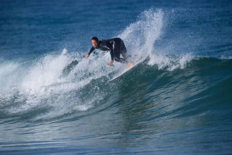 En surfer i en sort våddragt rider dygtigt på en bølge og skærer sig gennem vandet med en dynamisk spray. Havet er pulserende blåt under en klar himmel, der fanger øjeblikkets handling og energi.