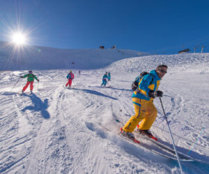 En gruppe skiløbere, iført farverige tøj, kører ned ad en sneklædt skråning under en klar blå himmel. Solen skinner klart og kaster lange skygger på sneen. Stolelifte og en bjergtop er synlige i baggrunden.