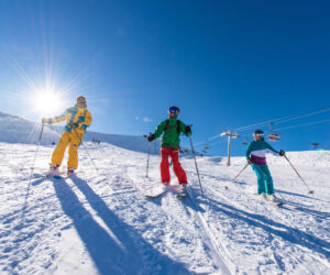 Tre skiløbere iført farverige tøj glider ned ad en sneklædt skråning under en klar blå himmel. Solen skinner klart og kaster skygger på sneen. Skilifte er synlige i baggrunden.
