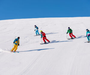 Fem personer på ski ned ad en sneklædt skråning på en klar solskinsdag. De er iført farverige skitøj inklusive gule, røde, grønne og blå jakkesæt. Himlen er lyseblå, og sneen er uberørt hvid.