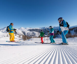 En gruppe på fire personer, der står på ski på en nypræpareret sneklædt skråning under en klar blå himmel. Tre står på ski ned ad bakke, mens en, iført knaldgul, står og ser på. Snedækkede bjerge er synlige i baggrunden.