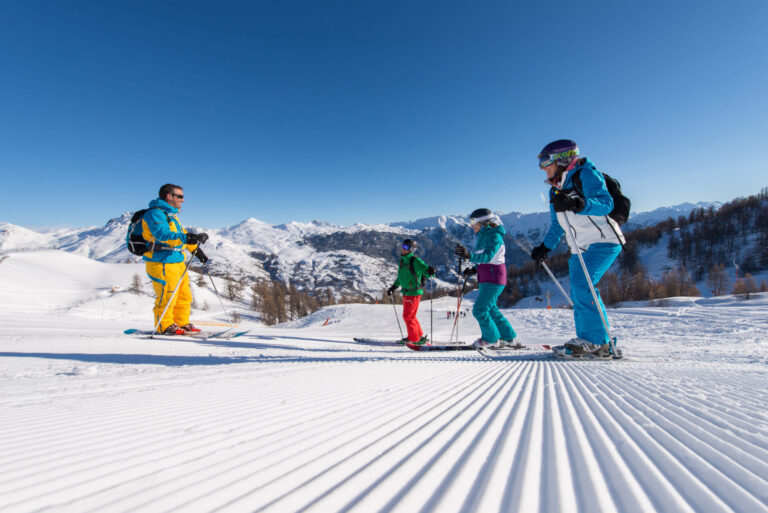 En gruppe på fire personer, der står på ski på en nypræpareret sneklædt skråning under en klar blå himmel. Tre står på ski ned ad bakke, mens en, iført knaldgul, står og ser på. Snedækkede bjerge er synlige i baggrunden.