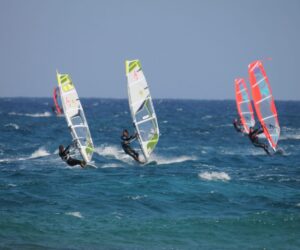 Fire windsurfere glider hen over et pulserende blåt hav på en solskinsdag, og deres farverige sejl fanger vinden. Himlen er klar, og små bølger bølger omkring dem og skaber en dynamisk og livlig scene.