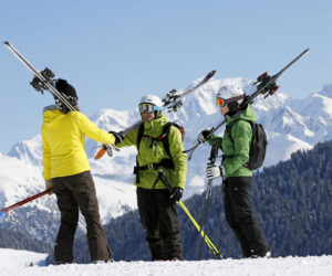 Tre skiløbere med skiudstyr slynget over skuldrene står på en sneklædt bjergskråning. De har farverige vinterjakker og hjelme på. Snedækkede tinder og en klar blå himmel ses i baggrunden.