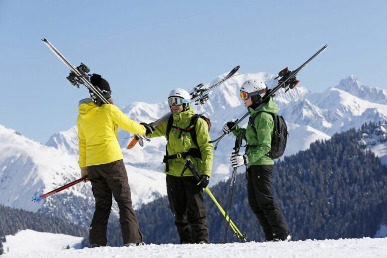 Tre skiløbere med skiudstyr slynget over skuldrene står på en sneklædt bjergskråning. De har farverige vinterjakker og hjelme på. Snedækkede tinder og en klar blå himmel ses i baggrunden.