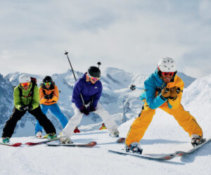 Fire skiløbere i farverige outfits og hjelme navigerer ned ad en sneklædt skråning med bjerge i baggrunden. De ser ud til at nyde vintersporten under en klar himmel.