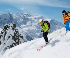 To skiløbere i vinterudstyr, den ene i grøn og den anden i orange, går ned ad en sneklædt bjergskråning. Majestætiske snedækkede tinder og delvist skyet blå himmel fungerer som baggrund. Skråningen er stejl, hvilket indikerer en spændende tur ned ad bakke.