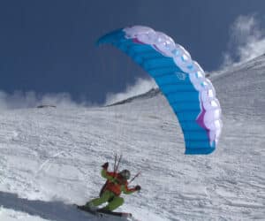 En person står på ski ned ad bakke på en sneklædt skråning, mens han betjener en blå faldskærm på den klare himmel og deltager i fartridning. Scenen er sat på baggrund af lys sne og en dybblå himmel.