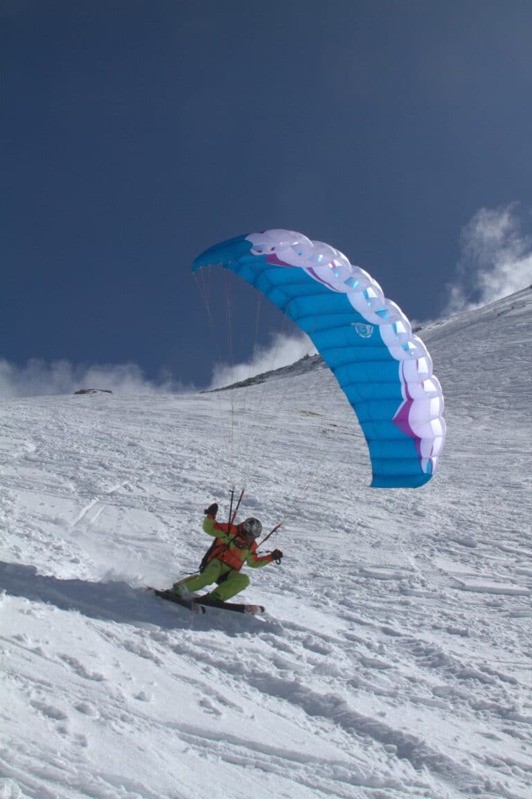En person står på ski ned ad bakke på en sneklædt skråning, mens han betjener en blå faldskærm på den klare himmel og deltager i fartridning. Scenen er sat på baggrund af lys sne og en dybblå himmel.