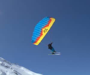 En person er paragliding, mens han står på ski ned ad en sneklædt skråning. Faldskærmen er farvestrålende i blå, gul og rød. Himlen er klar og blå, hvilket skaber en slående kontrast til den hvide sne.