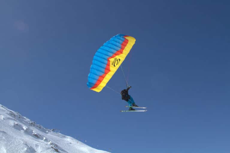 En person er paragliding, mens han står på ski ned ad en sneklædt skråning. Faldskærmen er farvestrålende i blå, gul og rød. Himlen er klar og blå, hvilket skaber en slående kontrast til den hvide sne.