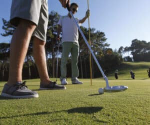 To golfspillere på en solrig bane med fokus på et putt. Golfspilleren i forgrunden har shorts og bådsko på, mens den anden holder flagstangen. Træer og andre spillere er synlige i baggrunden.