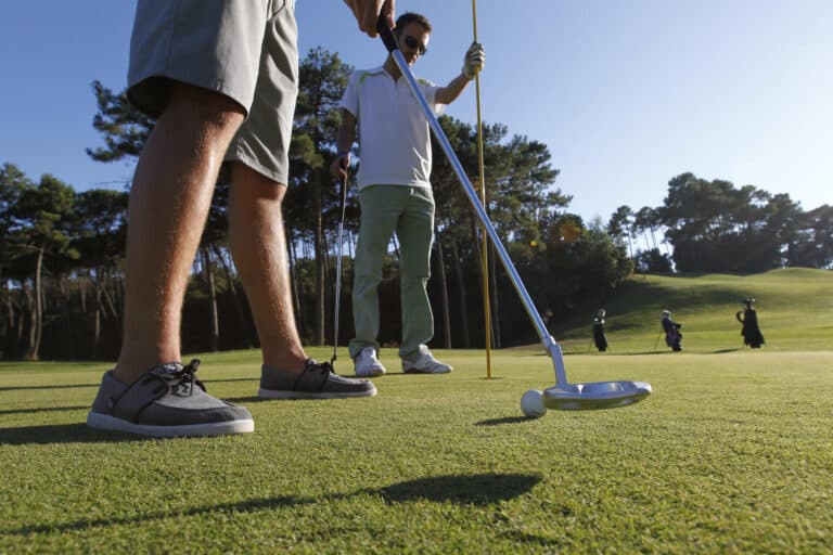 To golfspillere på en solrig bane med fokus på et putt. Golfspilleren i forgrunden har shorts og bådsko på, mens den anden holder flagstangen. Træer og andre spillere er synlige i baggrunden.