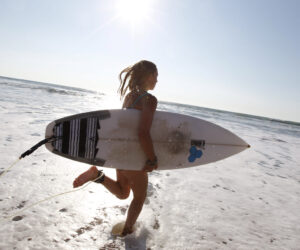En person med et surfbræt under armen går ud i havet. Solen skinner klart på himlen og kaster refleksioner på vandet. Scenen fanger et øjebliks forventning og spænding før surfing.