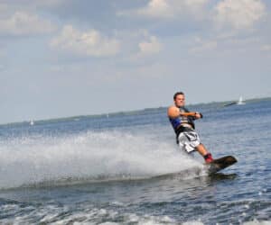 En person wakeboarder på en stor vandmasse og skaber en vandsprøjt bag ham. De er iført blå redningsvest og shorts. Himlen er delvist overskyet, og et par sejlbåde er synlige i det fjerne.