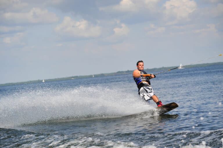 En person wakeboarder på en stor vandmasse og skaber en vandsprøjt bag ham. De er iført blå redningsvest og shorts. Himlen er delvist overskyet, og et par sejlbåde er synlige i det fjerne.