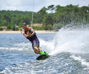 En person, der wakeboarder på en sø, læner sig til siden med vandsprøjt. De er iført en lys våddragt og grønne støvler med en skovklædt kyst i baggrunden under en delvist overskyet himmel.