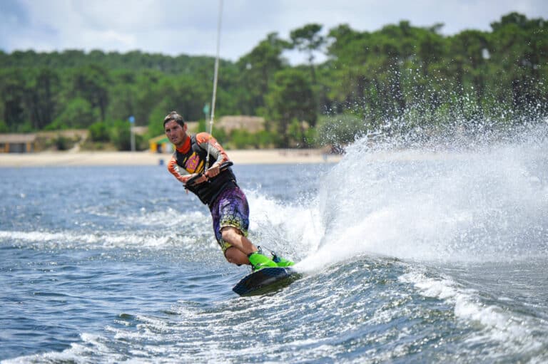 En person, der wakeboarder på en sø, læner sig til siden med vandsprøjt. De er iført en lys våddragt og grønne støvler med en skovklædt kyst i baggrunden under en delvist overskyet himmel.