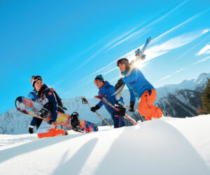 Tre personer i lyst vinterudstyr smiler og sludrer på et sneklædt bjerg. En holder et snowboard, og en anden bærer ski. Solen skinner klart over maleriske bjergtoppe i baggrunden.