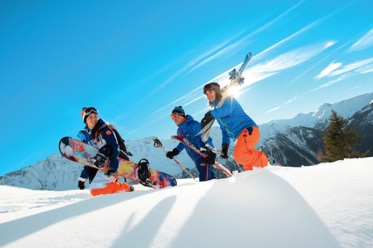 Tre personer i lyst vinterudstyr smiler og sludrer på et sneklædt bjerg. En holder et snowboard, og en anden bærer ski. Solen skinner klart over maleriske bjergtoppe i baggrunden.